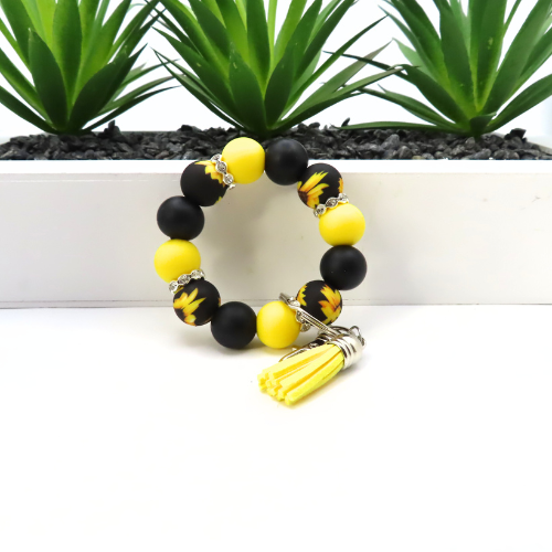 Black and yellow beaded bracelet with a tassel on a white surface with green plants in the background