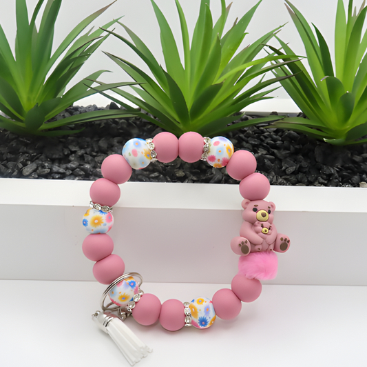 Pink beaded bracelet with teddy bear charm on a white surface with plants in the background