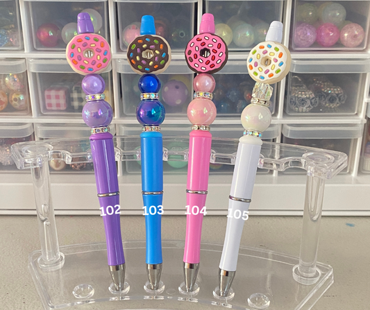 Four colorful pens with decorative beads on a clear stand, with storage bins in the background.