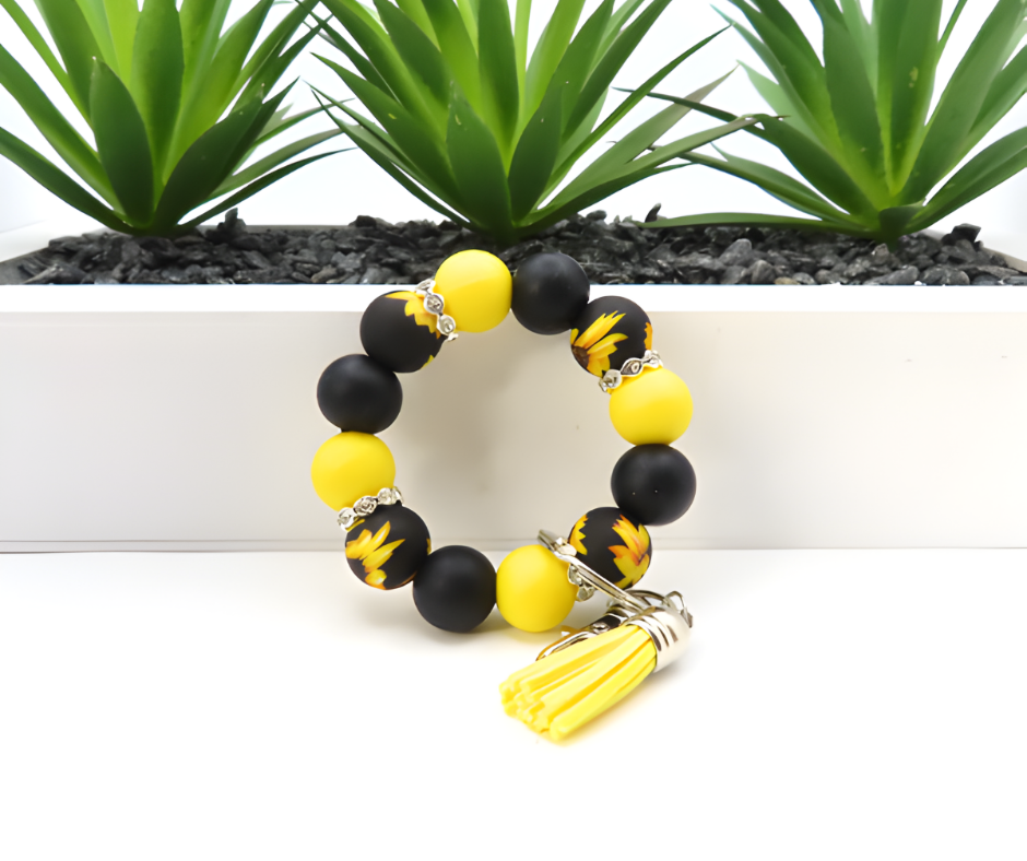 Black and yellow beaded bracelet with a tassel charm on a white surface with green plants in the background.