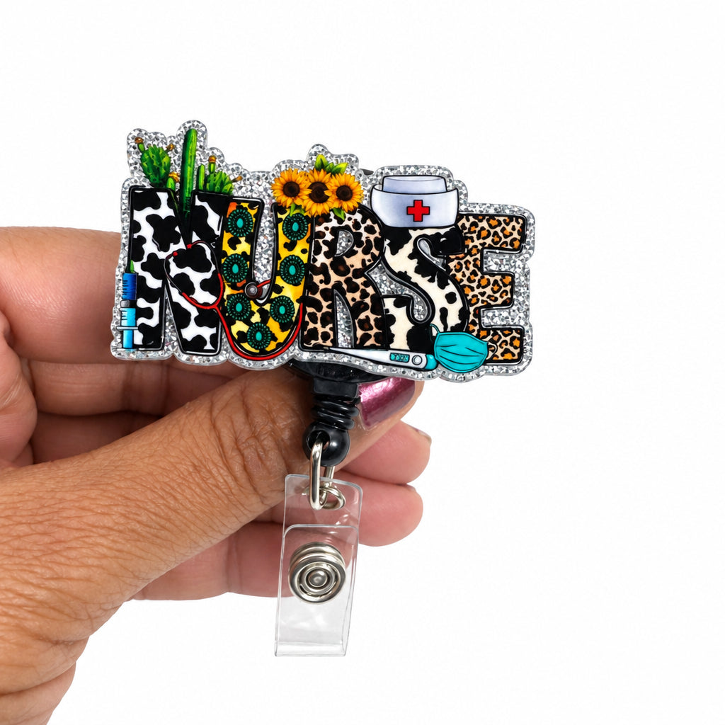 A hand holding a custom enamel badge reel with a decorative 'Nurse' design featuring leopard print, cow print, a stethoscope, a syringe, and a clipboard on a white background.
