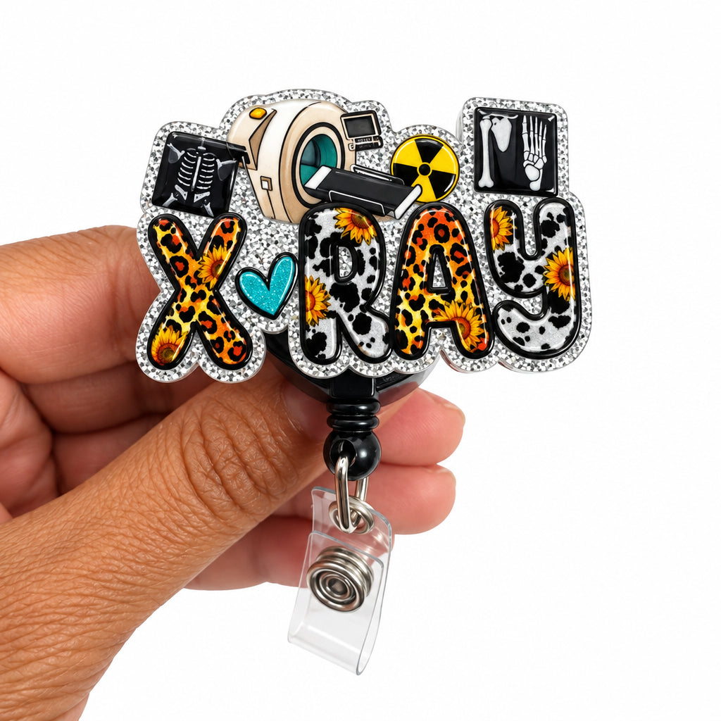 A hand holding a custom rhinestone-embellished 'X-Ray' badge reel. The letters feature leopard print, black-and-white cat patterns, a small turquoise heart, and a cartoon X-ray film icon.