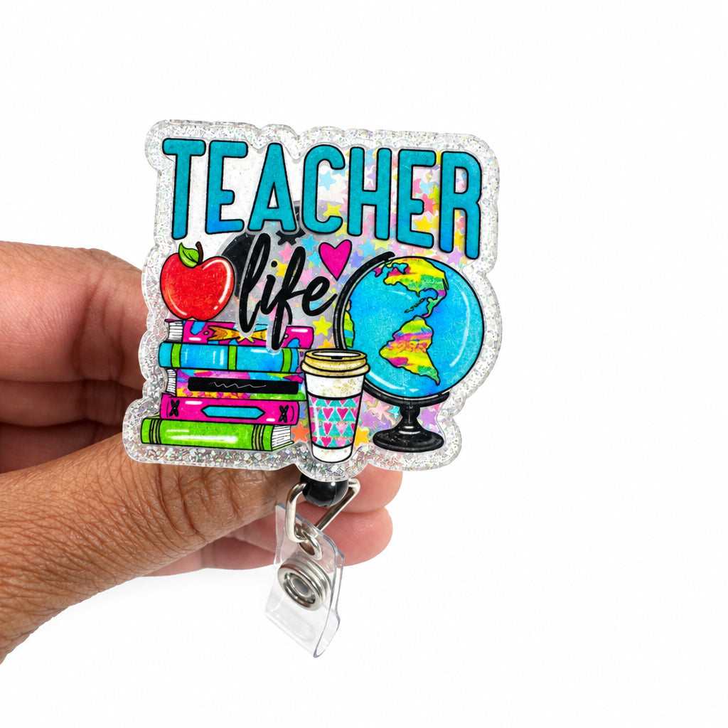 A hand holding a clear acrylic retractable badge reel keychain featuring a colorful 'TEACHER' design with school-themed illustrations like an apple, books, a globe, and a coffee cup, against a plain white background.