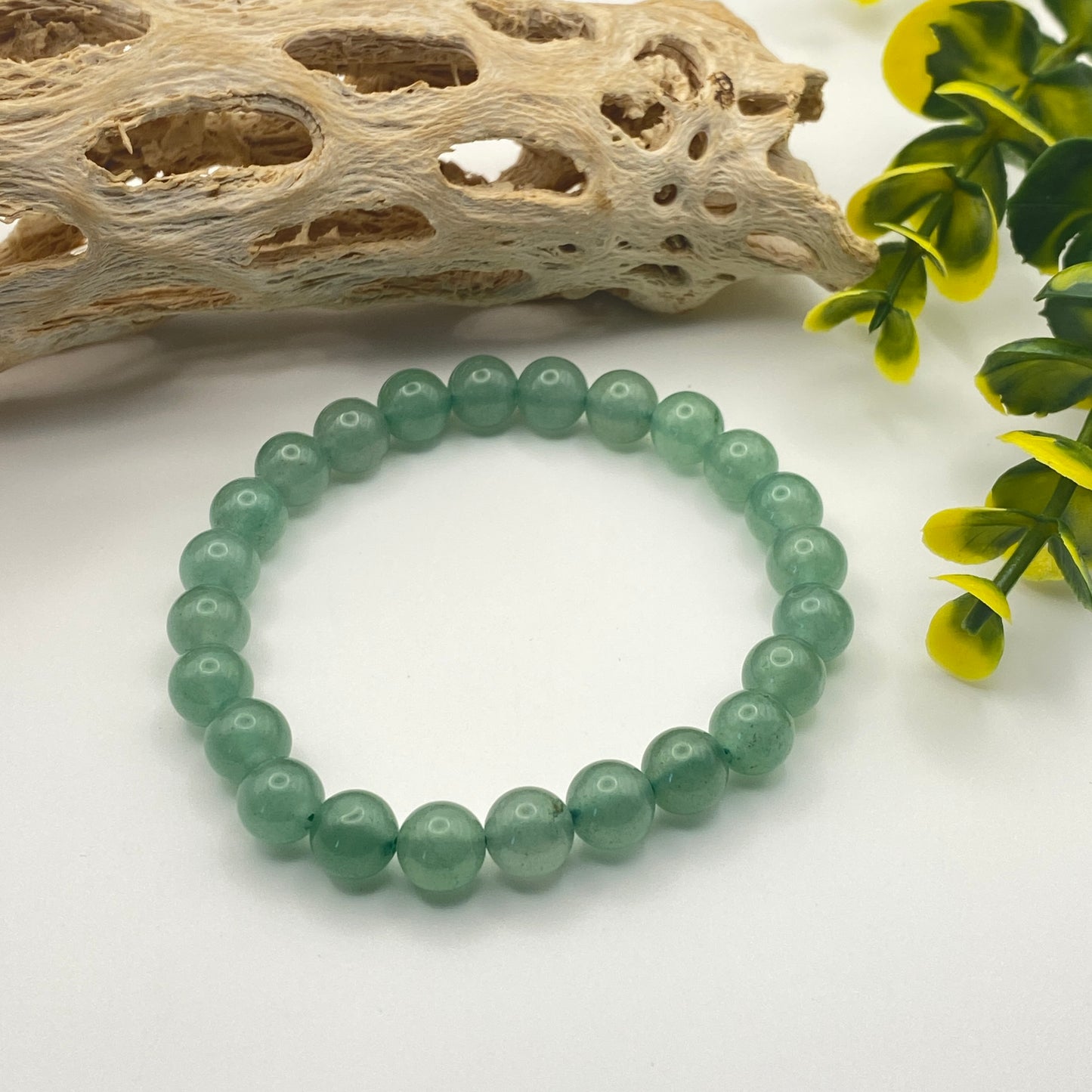 Green beaded bracelet on a white surface with natural elements like driftwood and leaves.