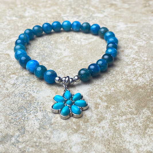 Blue beaded bracelet with a silver and turquoise flower charm on a textured beige surface