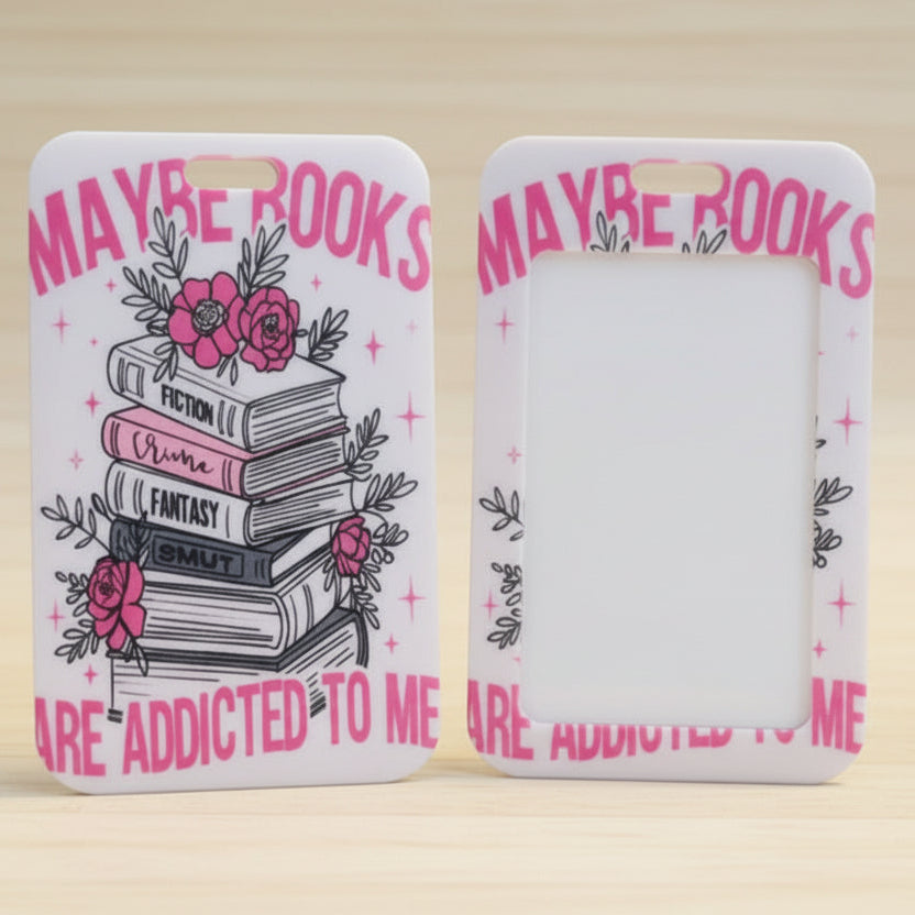 Two views of a white phone case with a pink and black book-themed design, standing on a light wood surface. The left case shows a stack of books with pink roses and the text 'MAYBE BOOKS ARE ADDICTED TO ME', while the right case shows a blank white back.