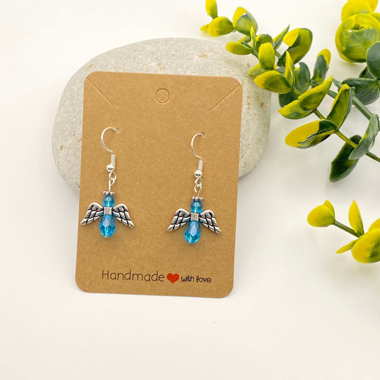 Pair of angel wing earrings with blue beads on a 'Handmade with Love' card, surrounded by greenery.