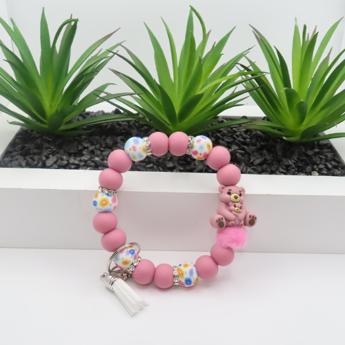 Pink beaded bracelet with teddy bear charm on a white surface with plants in the background