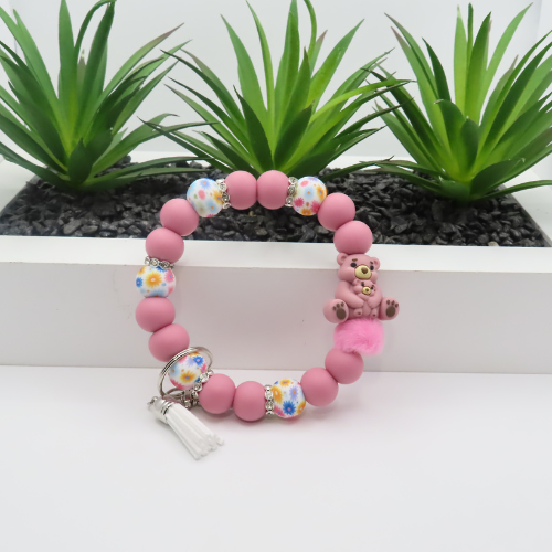 Pink beaded bracelet with floral charms and a white tassel on a white surface with plants in the background.