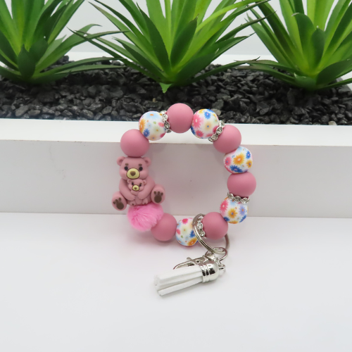Pink beaded bracelet with teddy bear charm and keychain on a white surface with green plants in the background