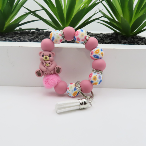 Pink beaded bracelet with a teddy bear charm and tassel on a white surface with plants in the background.