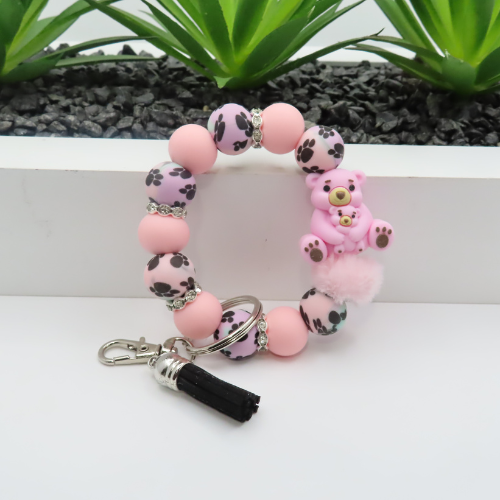 Pink paw print bracelet with a teddy bear charm on a white surface with green plants in the background
