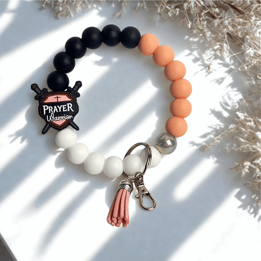 Beaded bracelet with a 'Prayer Warrior' charm on a white surface with dried plants.