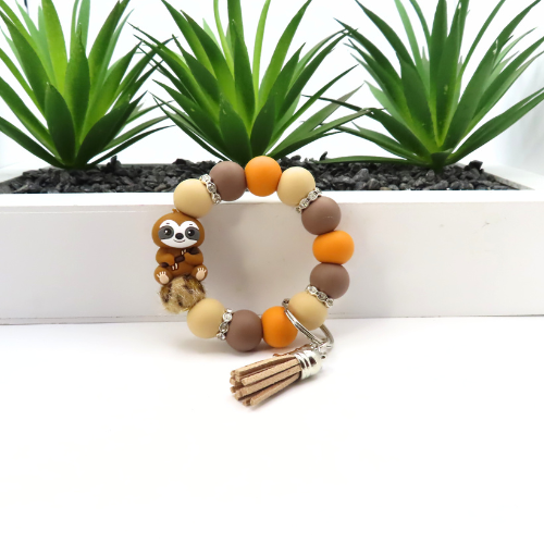 Beaded bracelet with sloth charm and tassel on a white surface with plants in the background