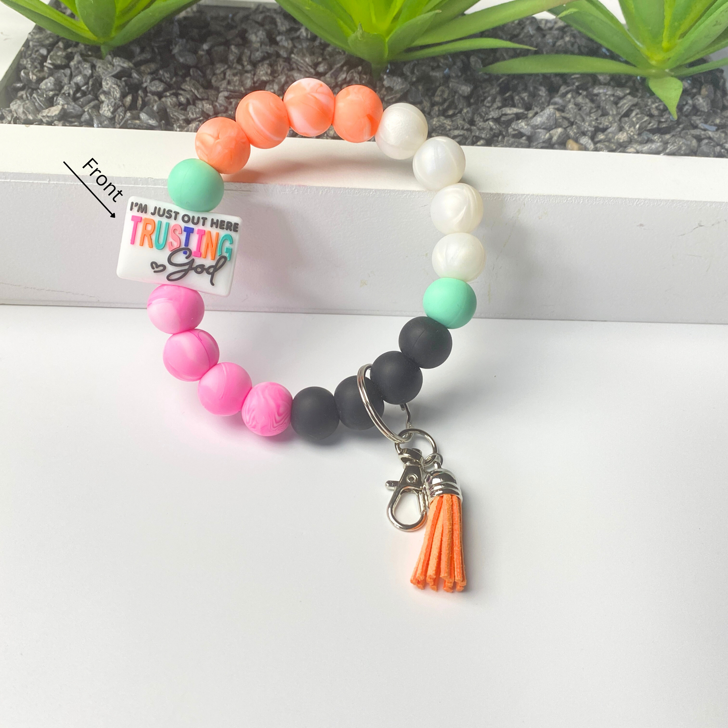 Colorful beaded bracelet with a tassel charm on a white surface, with a plant in the background.