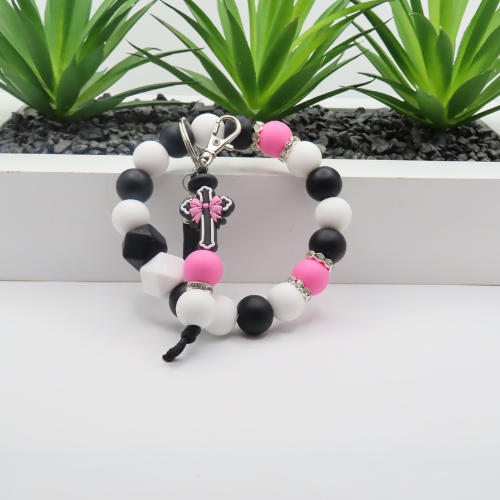 Beaded bracelet with a cross charm featuring pink accents on a white surface with green plants in the background.
