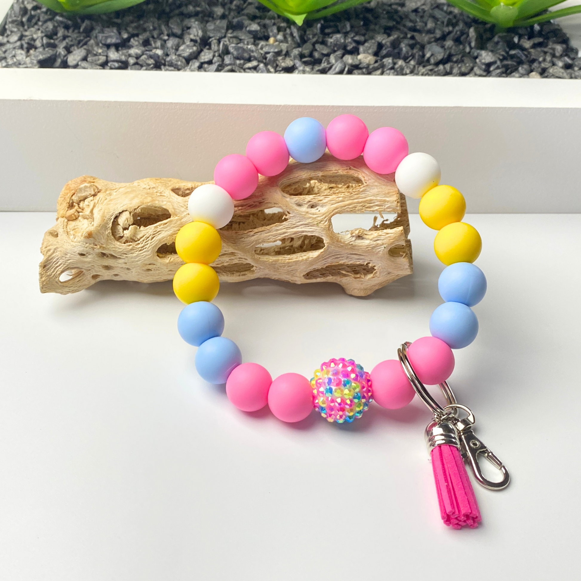 Colorful beaded bracelet with a keychain on a white surface with a natural wood piece and greenery in the background.