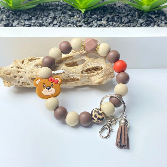 Beaded bracelet with a bear charm and keychain on a white surface with a natural wood piece and greenery in the background.