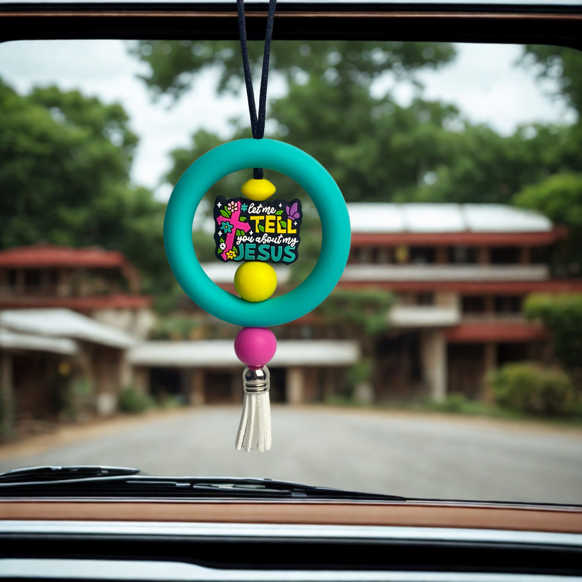 Colorful car hanging ornament with tassel on a car dashboard, blurred background of a building and trees.