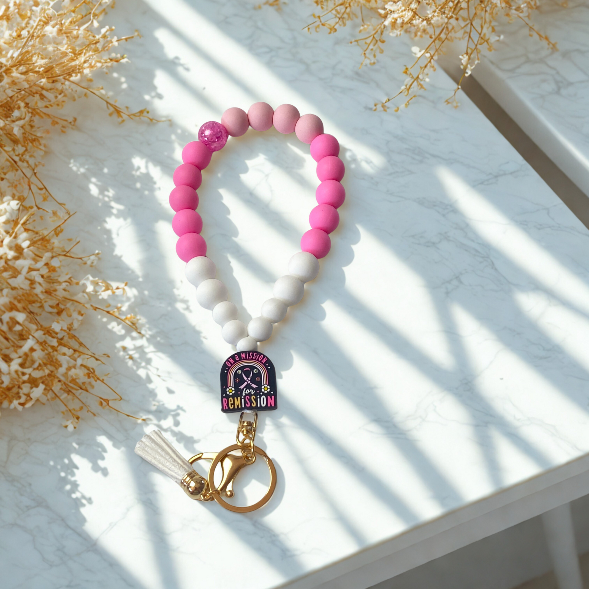 Pink beaded keychain with a gold ring and tassel on a white surface with decorative elements.