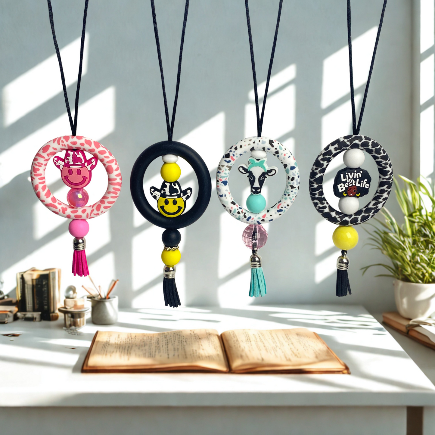 Four decorative necklaces with colorful beads and tassels on a desk with books and a plant.