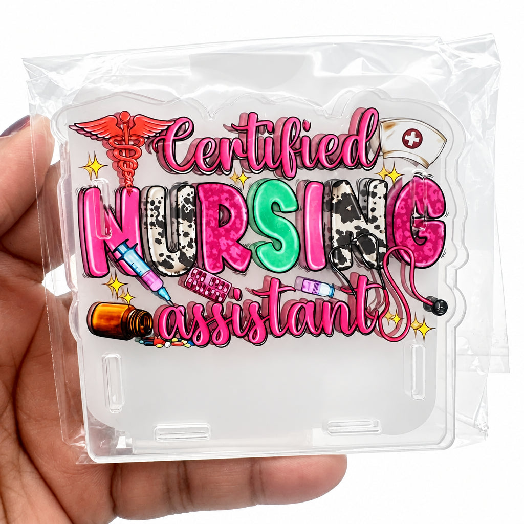A hand holding a clear plastic bag containing a decorative acrylic sign that reads 'Certified Nursing Assistant' in pink, green, and multicolor lettering with medical-themed illustrations.