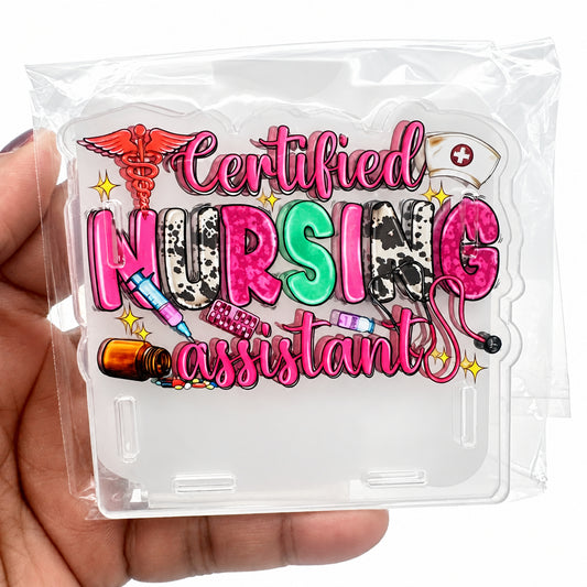 A hand holding a clear plastic bag containing a decorative acrylic sign that reads 'Certified Nursing Assistant' in pink, green, and multicolor lettering with medical-themed illustrations.