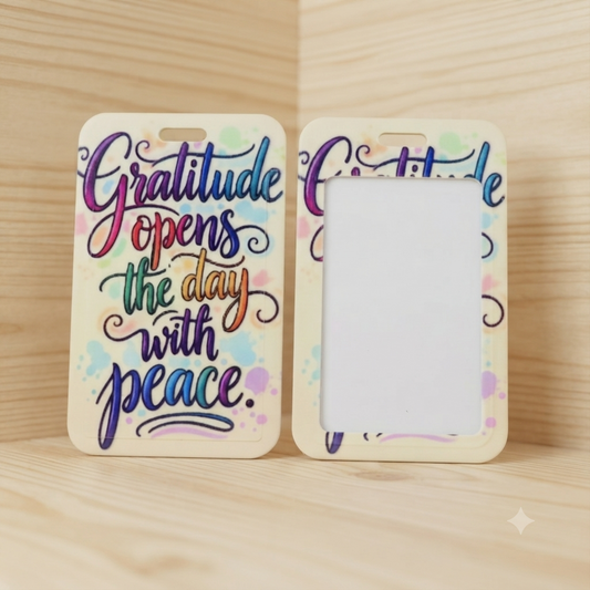 Two small rectangular card holders or frames with rounded corners, shown on a light wood surface. One has colorful hand-lettered text reading 'Gratitude opens the day with peace.