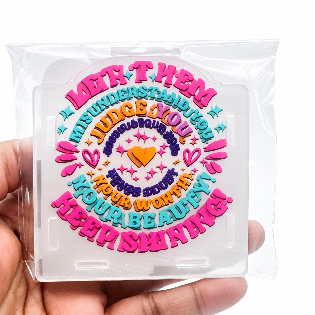 A hand holding a clear plastic case containing a colorful circular sticker or decal with decorative text and hearts. The design features pink, orange, purple, and blue lettering on a white background.
