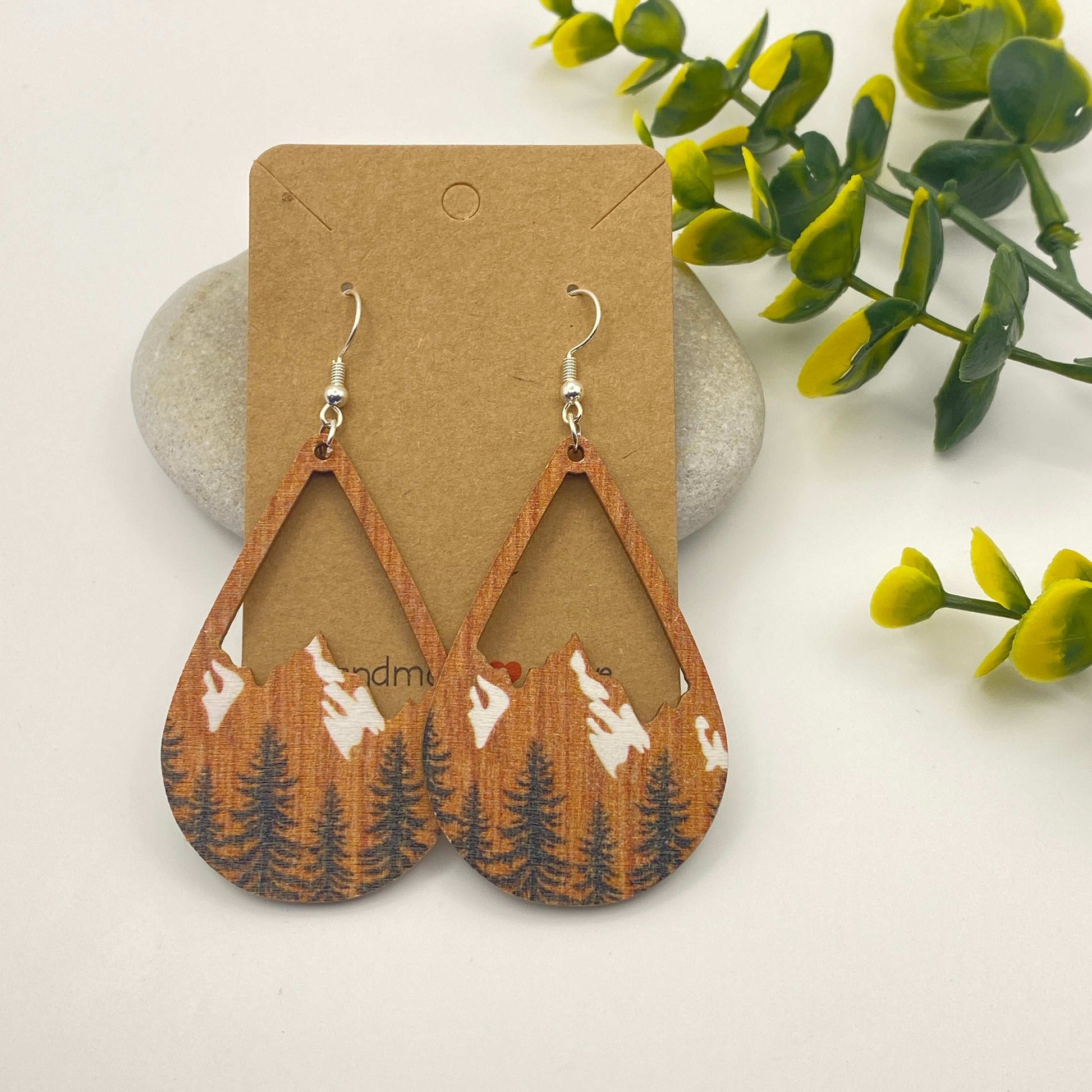 Wooden earrings with nature design on a cardboard backing, surrounded by greenery.