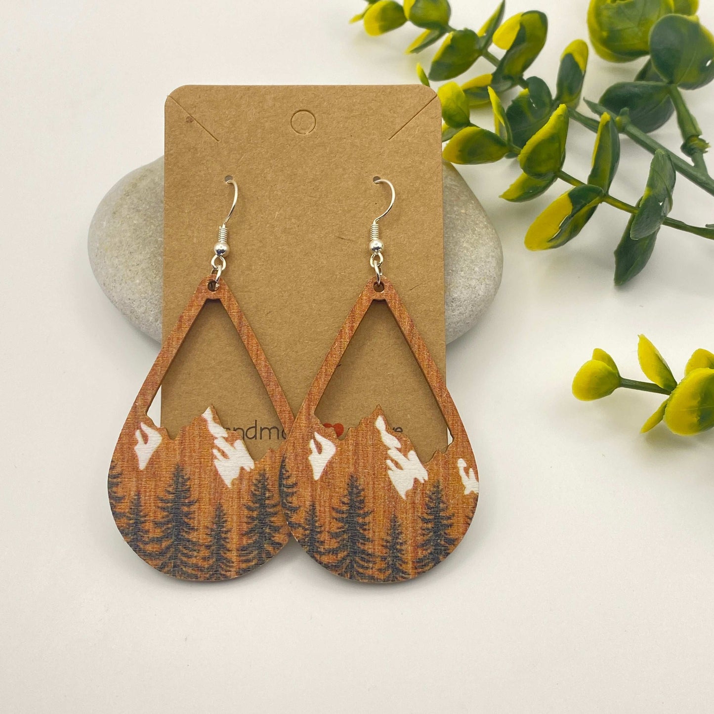 Wooden earrings with mountain and tree design on a cardboard backing, surrounded by green leaves.