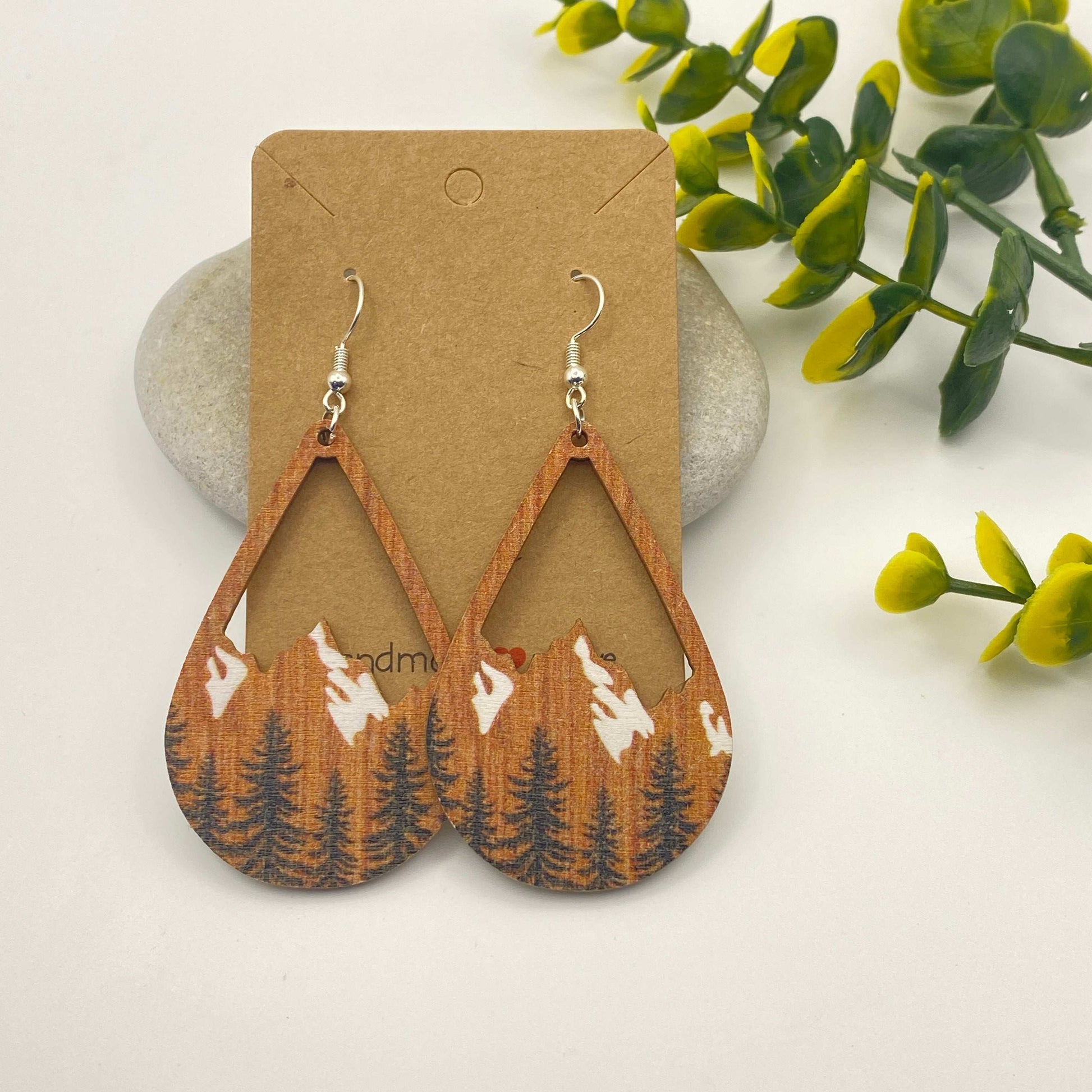 Wooden earrings with mountain and tree design on a cardboard backing, surrounded by green leaves.