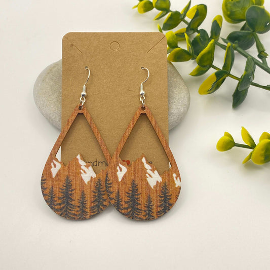Wooden earrings with mountain and tree design on a cardboard backing, surrounded by green leaves.