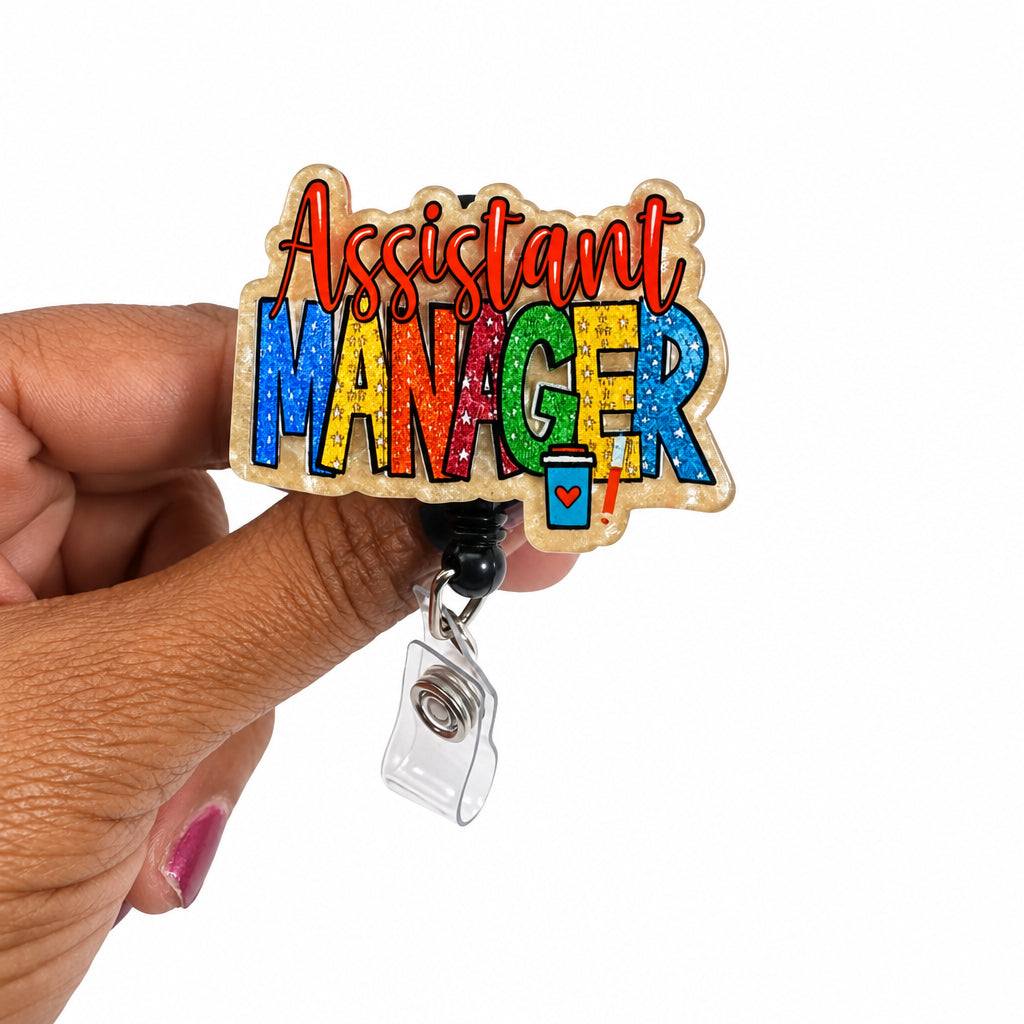 A hand holding a clear acrylic badge reel sign that reads 'Assistant Manager' in colorful glittery letters against a plain white background.