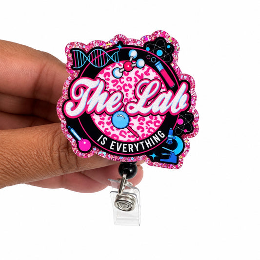 A hand holding a custom die-cut acrylic badge reel featuring a pink leopard print background, the text 'The Lab IS EVERYTHING' in white and black, and a black, pink, and blue retro sci-fi design with a holographic glitter border.