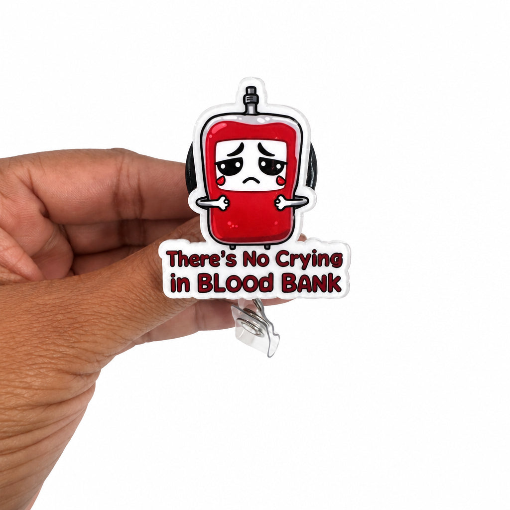 A hand holding a clear acrylic pin featuring a red blood bag character with a sad face and the text 'There's No Crying in BLOOD BANK' on a plain white background.