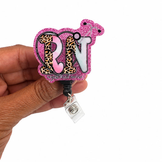 A hand holding a pink glitter and leopard print ID badge reel with the letters 'RN' in the center, set against a plain off-white background.