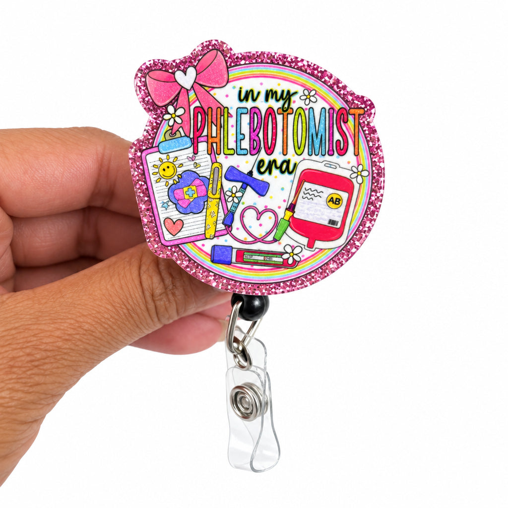 A hand holding a round pink glitter badge reel featuring a colorful illustration with the text 'In my PHLEBOTOMIST era!'.
