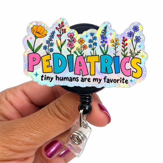 A hand holding a clear acrylic retractable badge reel featuring colorful floral artwork and the word 'PEDIATRICS' in pastel rainbow letters, with the phrase 'tiny humans are my favorite' below.