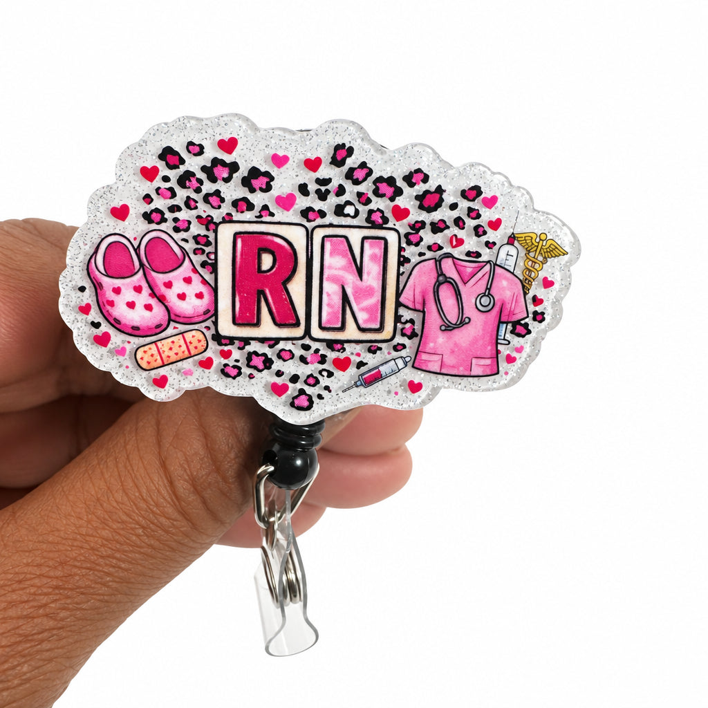 A hand holding a custom cloud-shaped acrylic badge reel cover featuring 'RN' lettering, pink shoes, pink scrubs, a stethoscope, and a syringe on a leopard print background with glitter.
