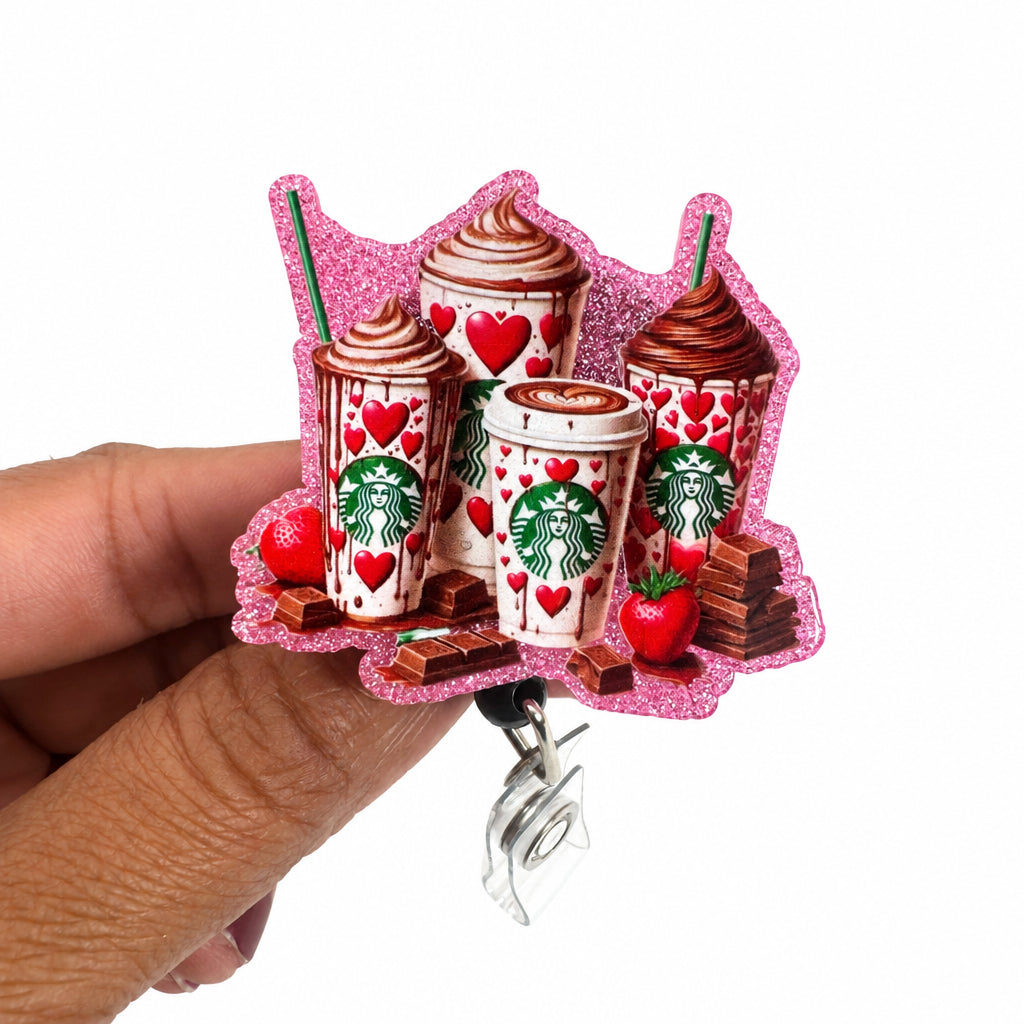 A hand holding a pink glitter charm featuring Starbucks-themed iced drinks with red hearts, strawberries, and chocolate pieces, with a silver metal clip attached at the bottom.