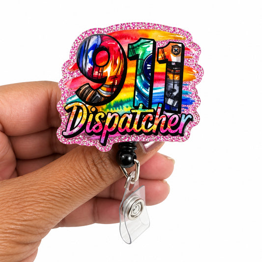 A hand holding a glittery acrylic badge reel featuring a colorful '911 Dispatcher' design with a rainbow gradient and holographic finish, set against a plain white background.