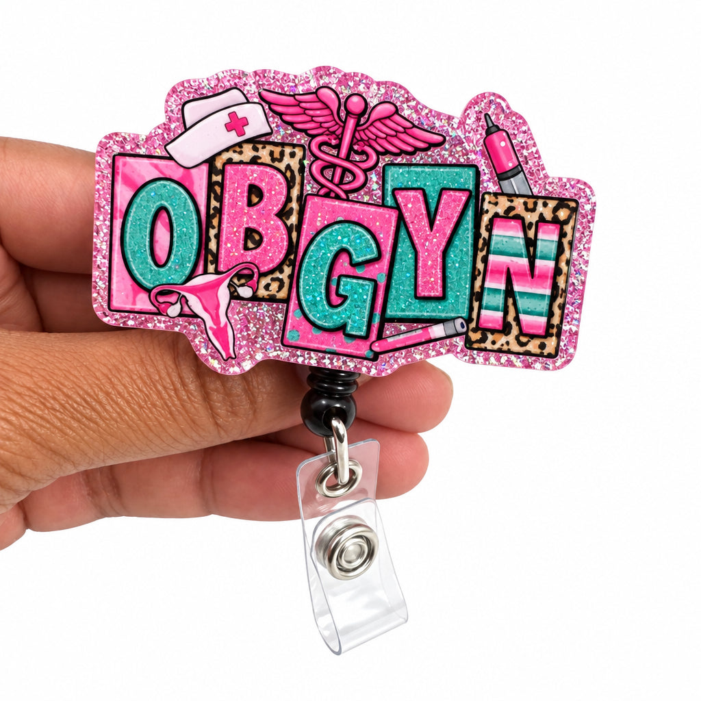 A hand holding a pink glitter 'OBGYN' badge reel with colorful letters and medical-themed designs, set against a plain white background.