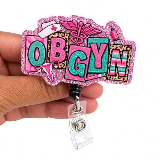 A hand holding a pink glitter 'OBGYN' badge reel with colorful letters and medical-themed designs, set against a plain white background.