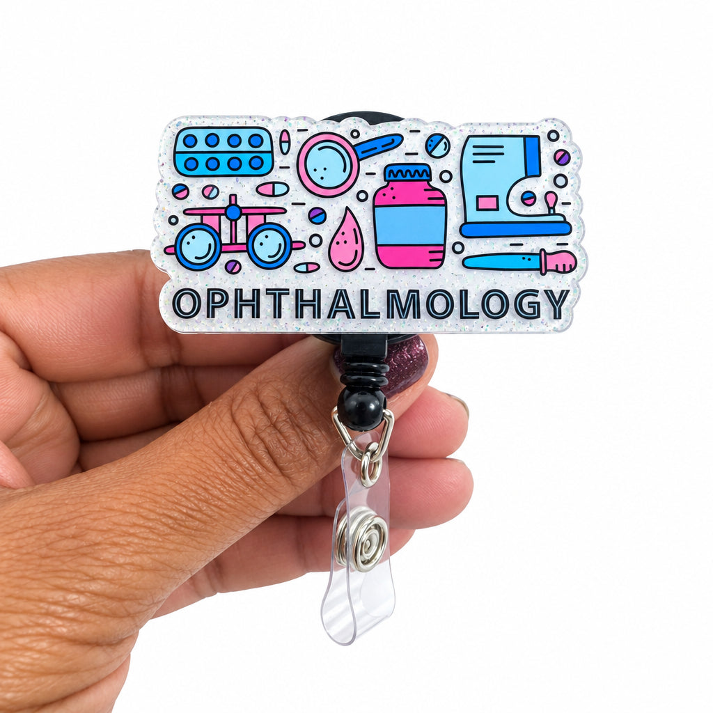A hand holding a clear acrylic retractable badge reel cover featuring colorful ophthalmology-themed illustrations and the word 'OPHTHALMOLOGY' in gray lettering, with a black and silver retractable strap below.