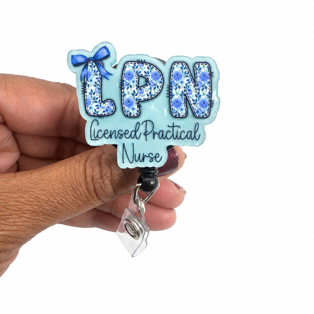 A hand holding a custom 'LPN Licensed Practical Nurse' badge reel. The badge has a light blue background with a blue floral pattern inside the letters 'LPN' and a small blue bow on top.