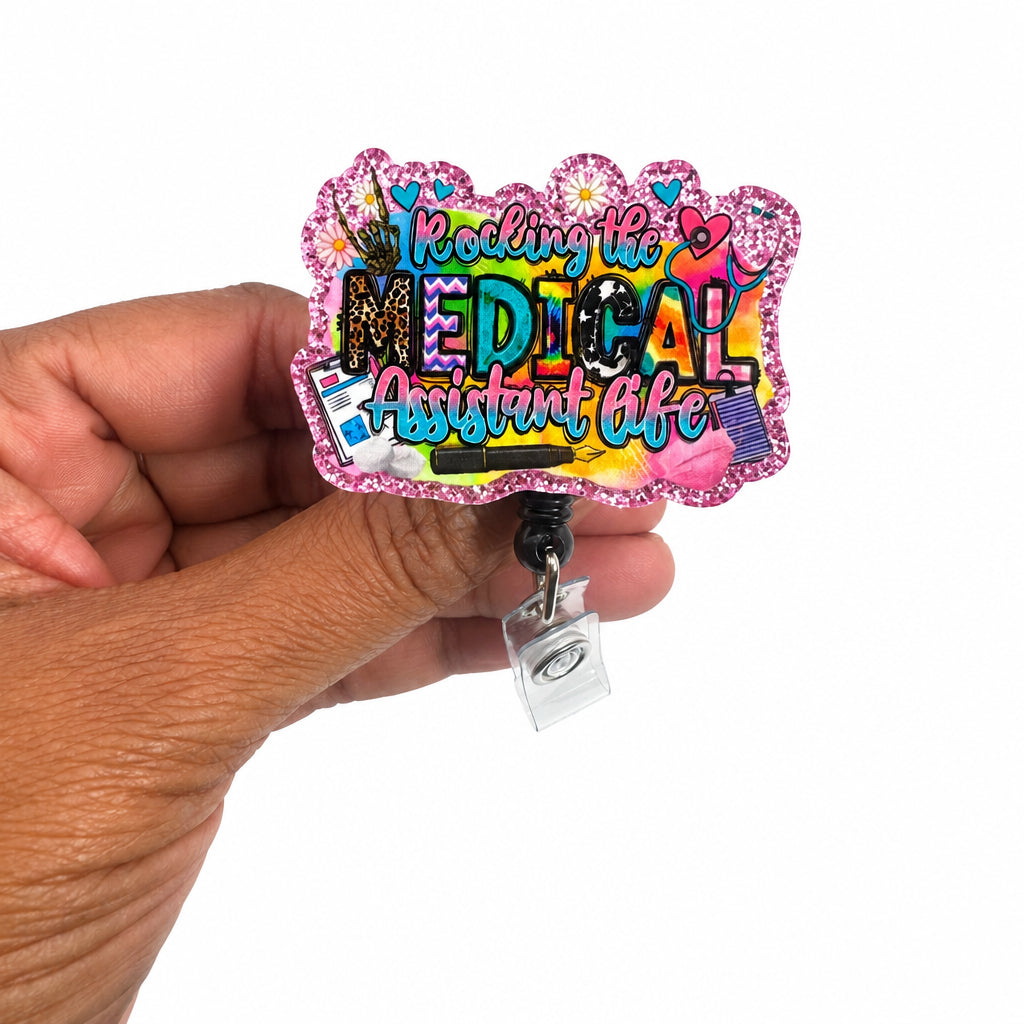 A hand holding a colorful, glittery acrylic badge reel. The badge reel features the text 'Reading the MEDICAL Assistant Life' in a vibrant, multicolored graffiti-style font with a pink glitter border.