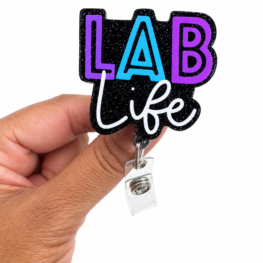A hand holding a die-cut acrylic badge reel featuring the text 'LAB life' in purple, light blue, and white script on a black glittery background, with a clear plastic ID holder attached.