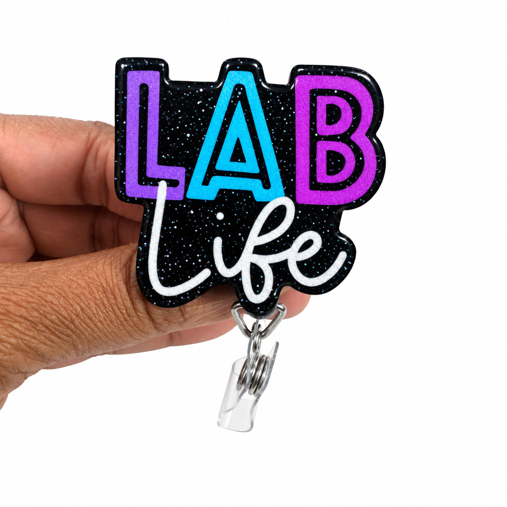 A hand holding a black acrylic badge reel cover featuring the text 'LAB life' in purple, blue, and white lettering against a black glittery background, with a clear plastic clip at the bottom.