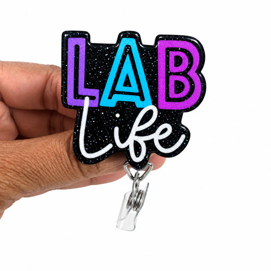 A hand holding a black acrylic badge reel cover featuring the text 'LAB life' in purple, blue, and white lettering against a black glittery background, with a clear plastic clip at the bottom.